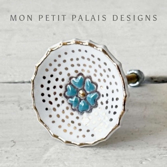 A white ceramic knob with a turquoise bloom center and gold edging.