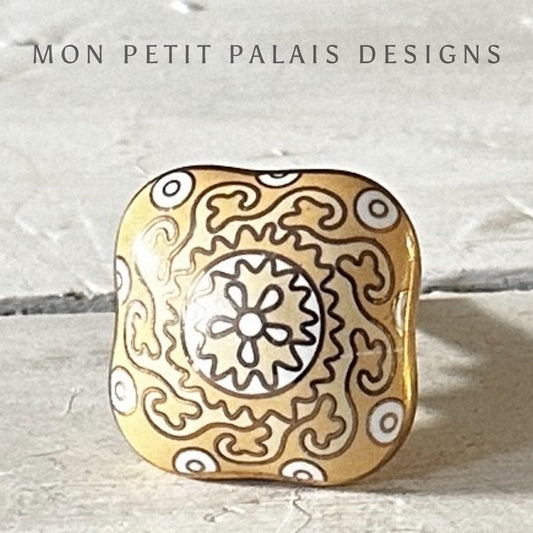 A gold-colored ceramic knob with a floral pattern, branded with 'MON PETIT PALAIS DESIGNS'.