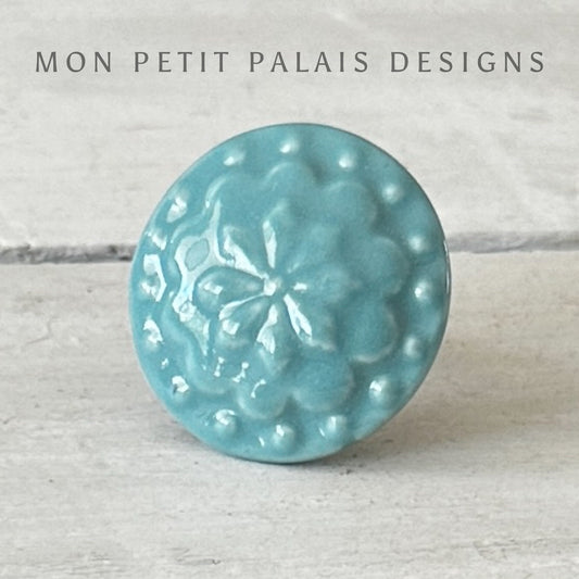 A turquoise ceramic knob with a floral design, branded with 'MON PETIT PALAIS DESIGNS'.