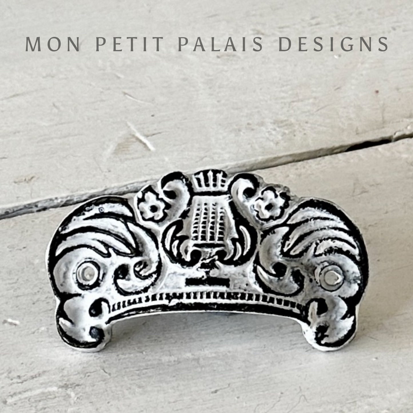 A white filigree metal cup handle with intricate designs, branded by MON PETIT PALAIS DESIGNS.