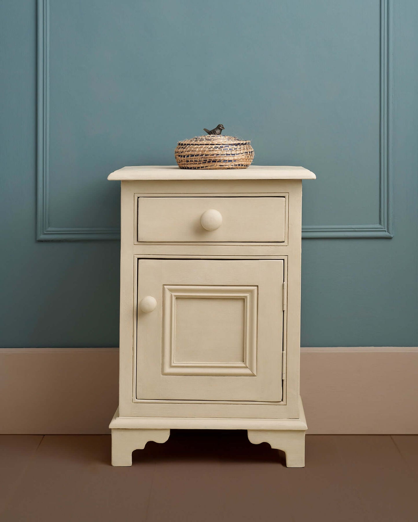 Bedside cabinet painted in Annie Sloan Canvas against a dark teal wall with a woven basket.