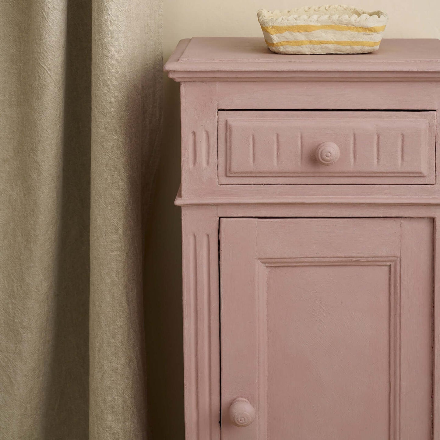 Close-up of a vintage bedside cabinet painted in Annie Sloan Jaipur Pink, styled against a neutral curtains.