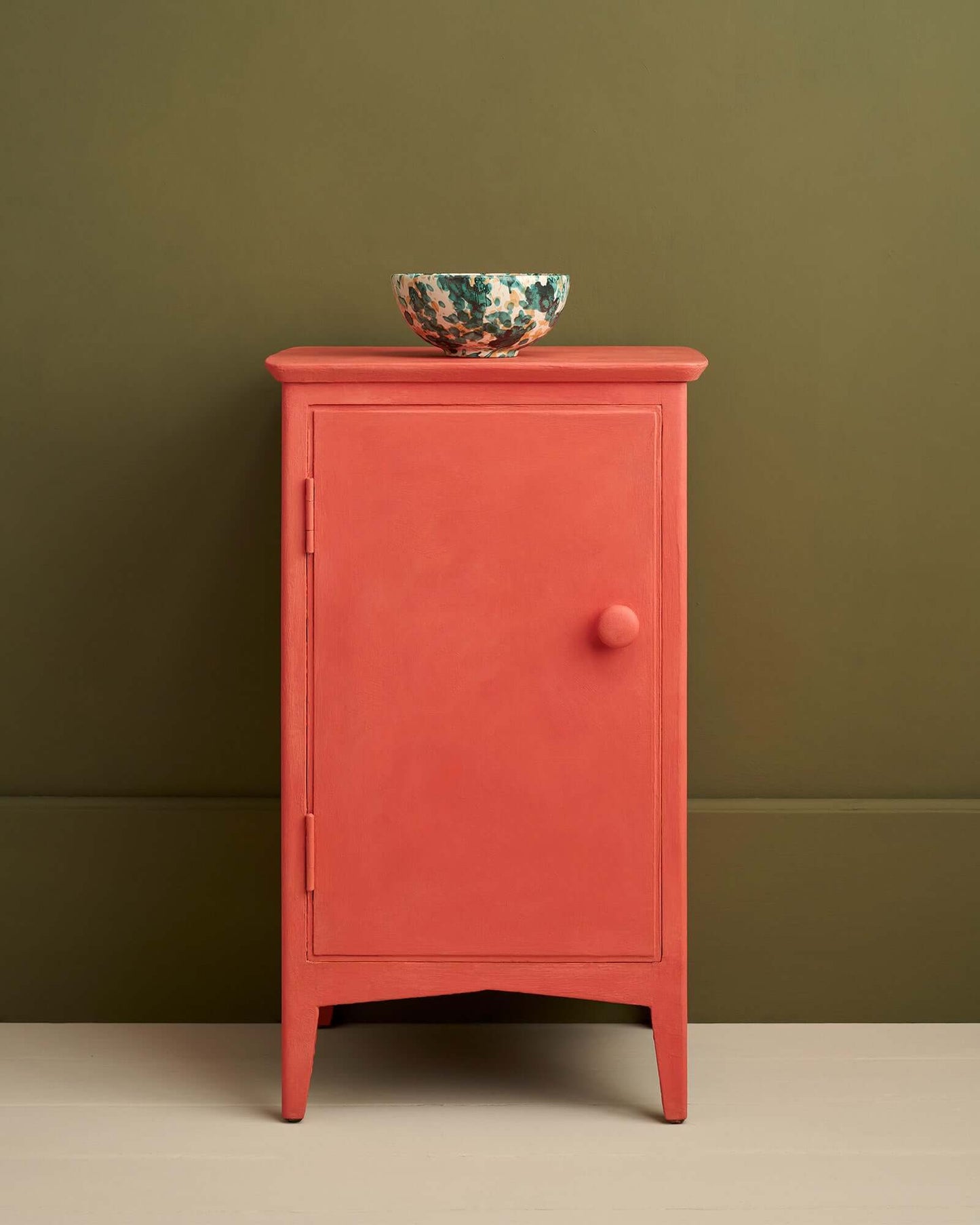 Small cabinet fully painted in Annie Sloan Sandalwood Red against an olive green wall, styled with a ceramic bowl.