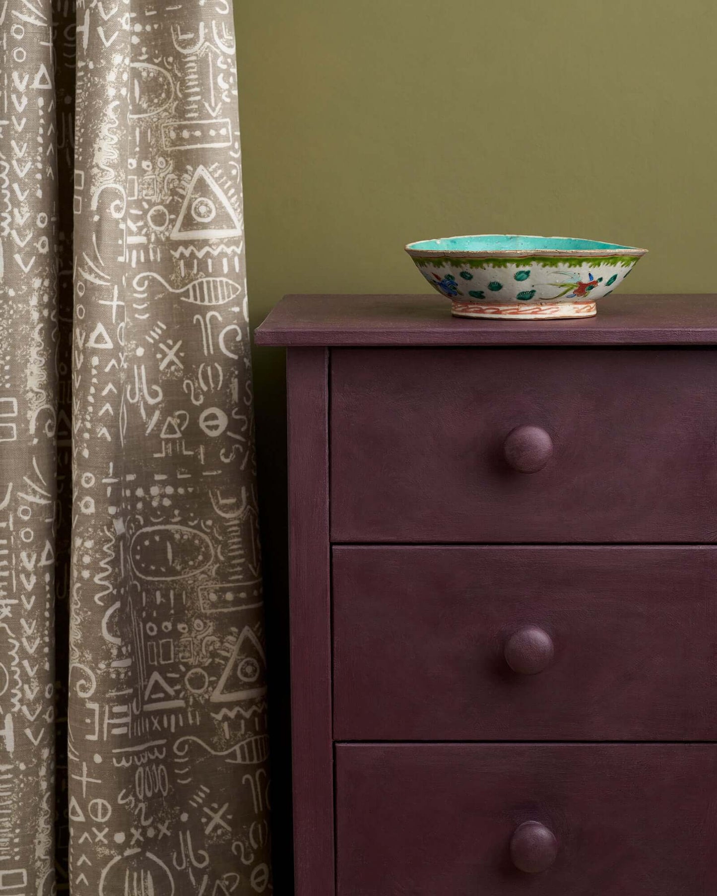 Close-up of a Tyrian Plum painted dresser with decorative bowl and patterned curtain against an olive green wall.