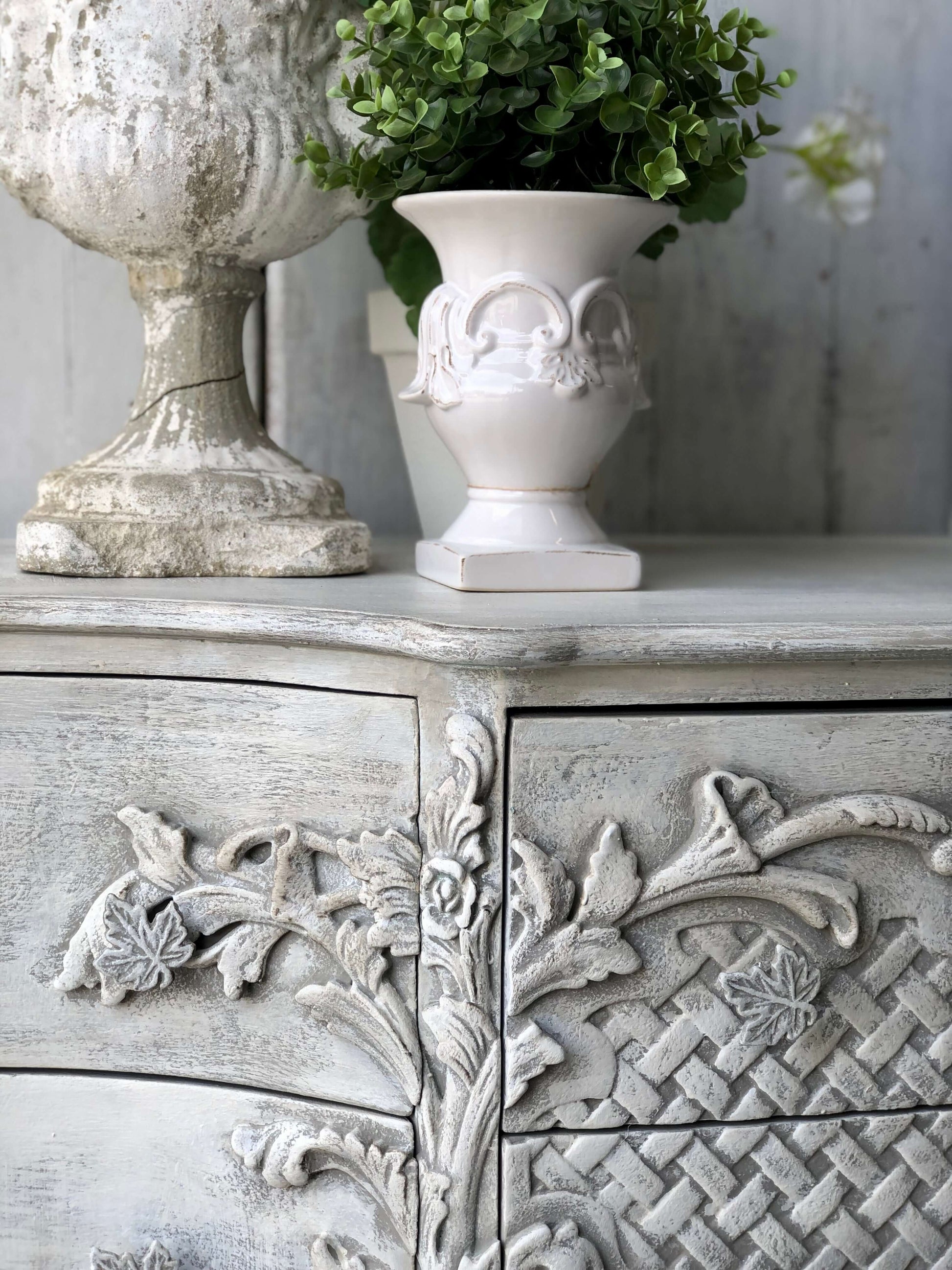 A close-up of an ornate cabinet with floral patterns, painted in a faded elegant gray color, with a decorative white vase on top.