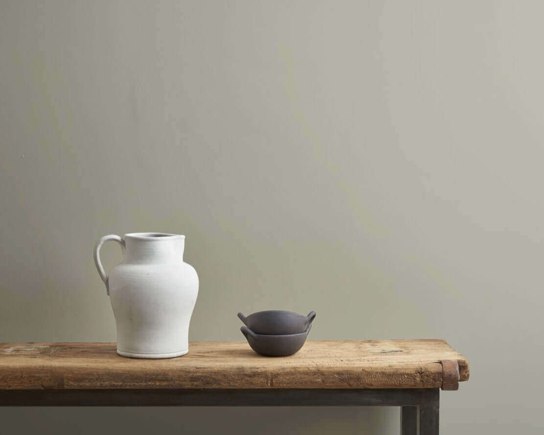 A white ceramic pitcher and a small black bowl on a wooden table against a soft green wall.