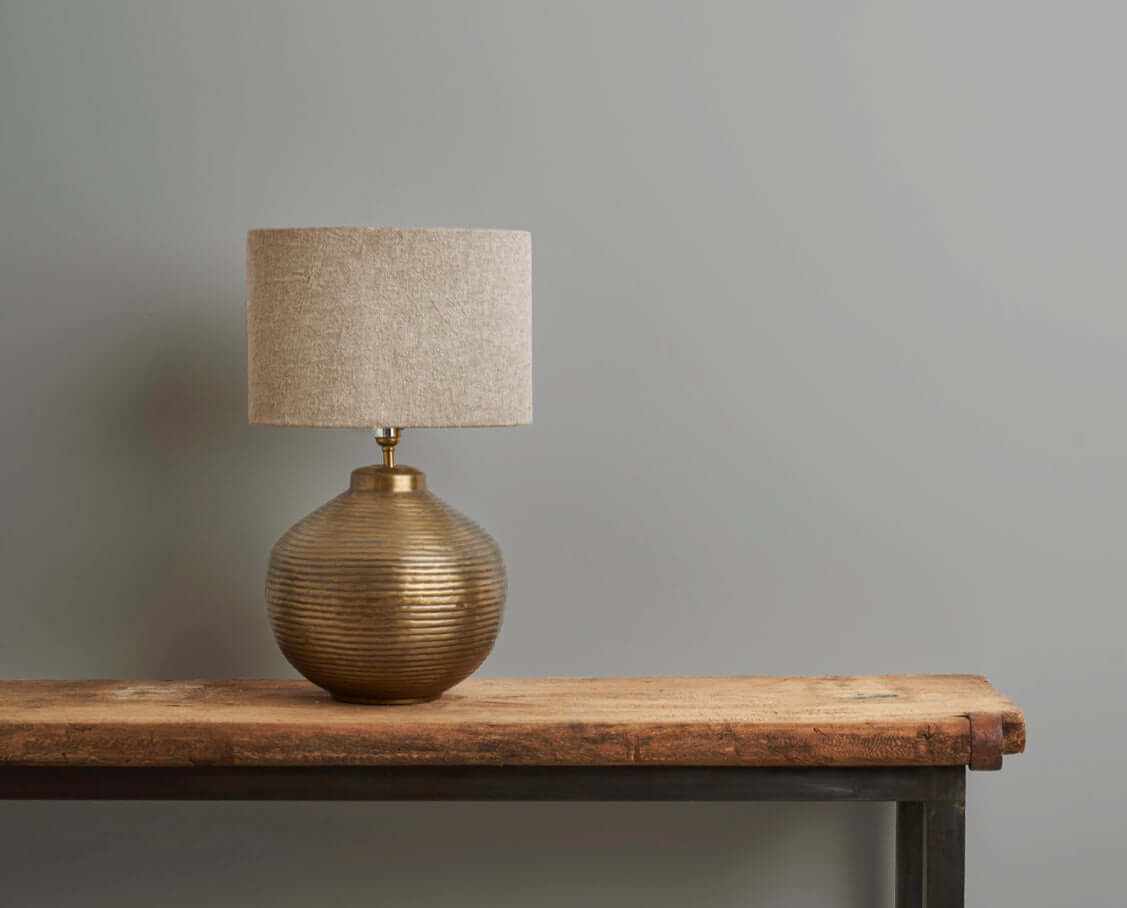 A decorative lamp with a ribbed gold base and a light fabric shade, placed on a wooden table against a grey wall.