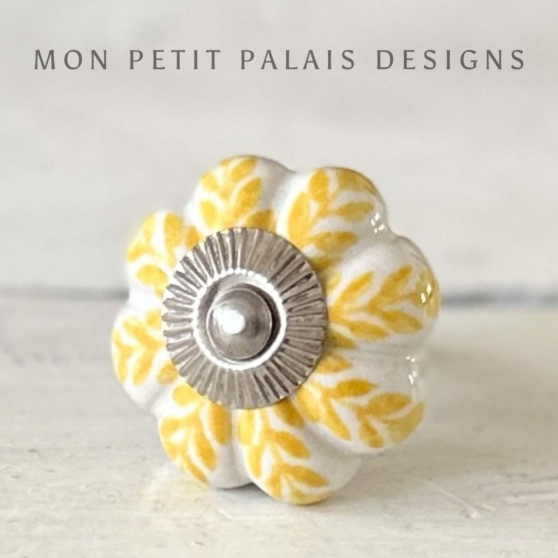 A round ceramic knob with a yellow leaf pattern and a central silver screw, against a white background.