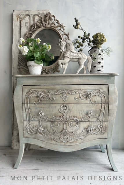 Decorative chest of drawers with ornate carvings, mirror, and plants in a room setting.