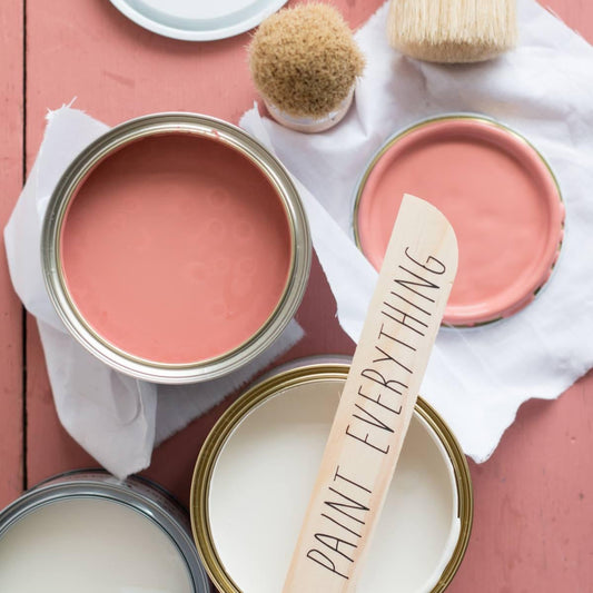 Four Annie Sloan Chalk paint cans with different colors and a paintbrush labeled 'Paint Everything' on a pink wooden surface.