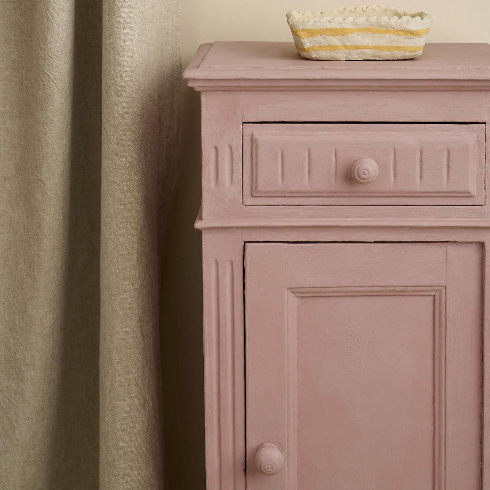 Close-up of a vintage bedside cabinet painted in Annie Sloan Jaipur Pink, styled against a neutral curtains. 
