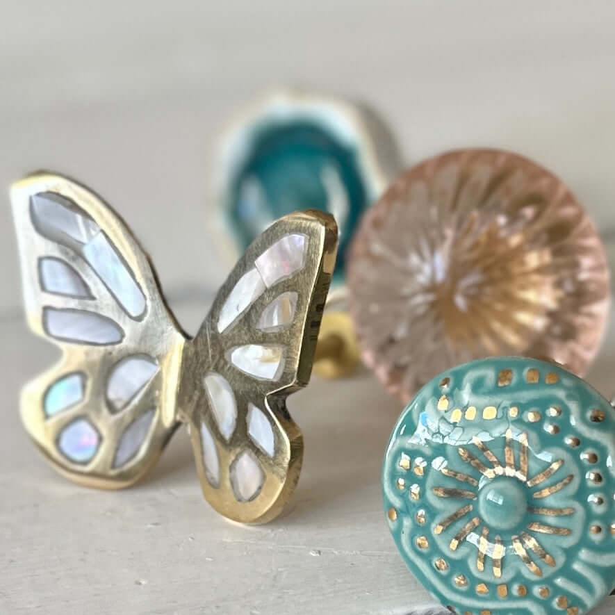 Butterfly brass & mother of pearl cabinet knob alongside decorative glass knobs on a white surface.