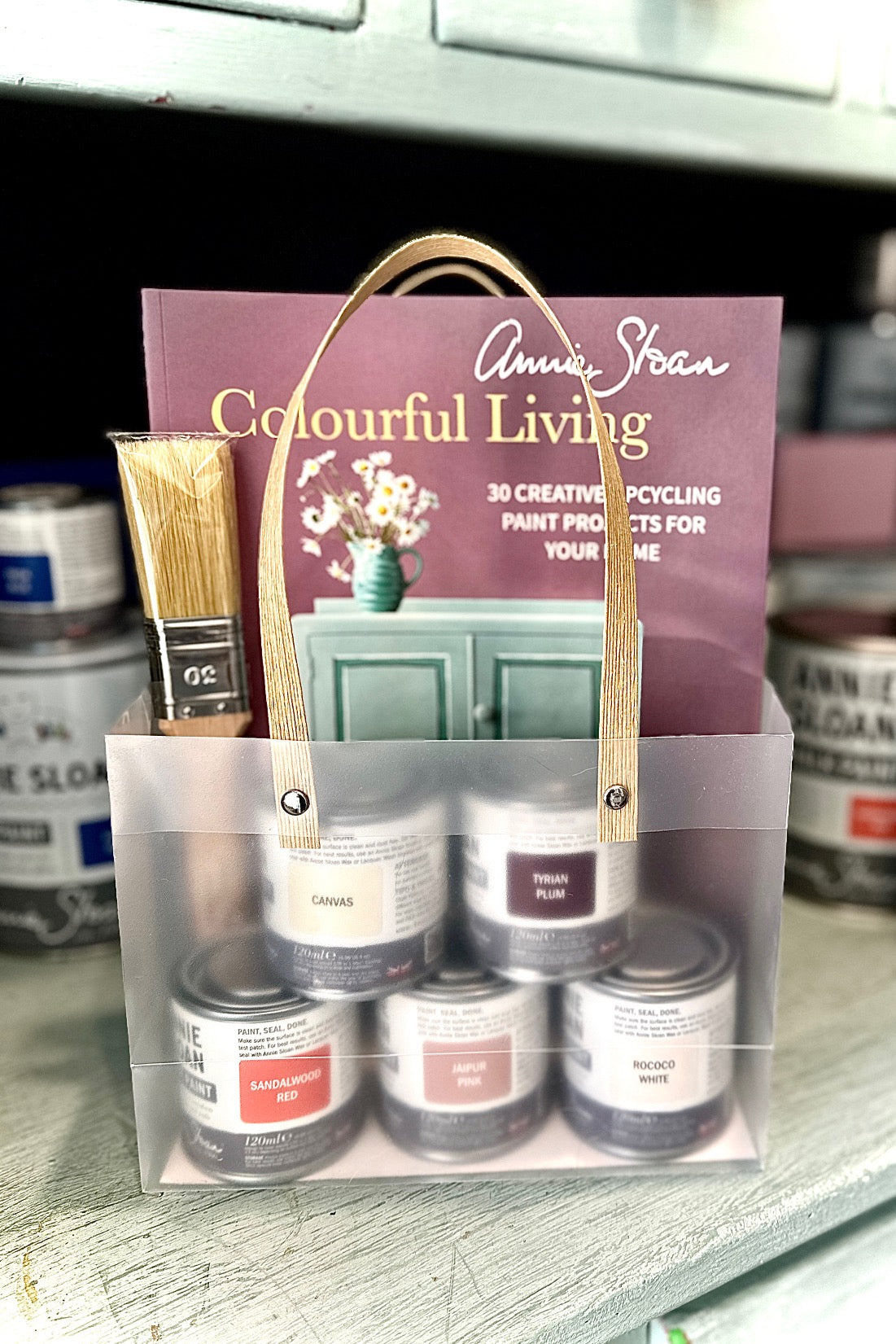 Set of paint cans with a brush and a book titled 'Colourful Living' by Annie Sloan on a wooden surface.