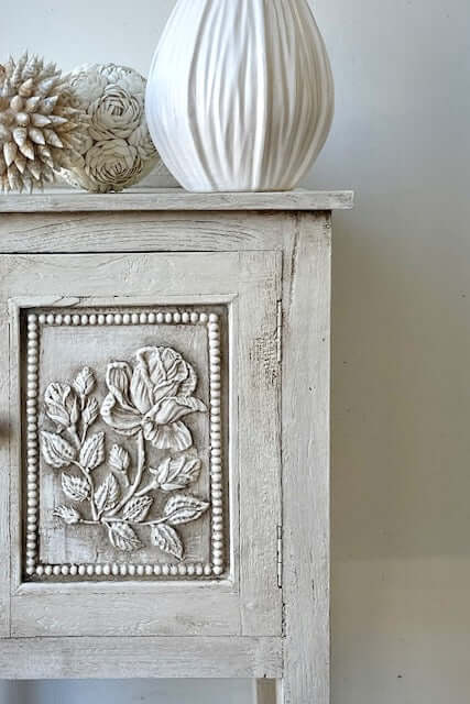 Wildflower cabinet with decorative wood detail and floral carvings and a white vase on top against a plain wall.