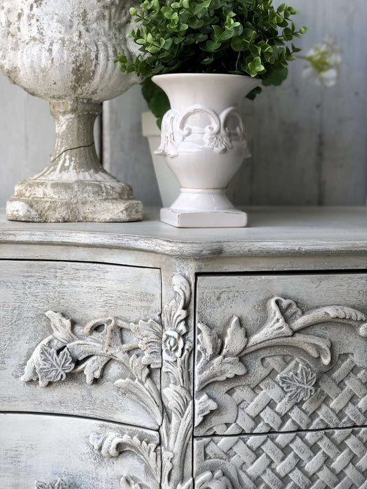 A close-up of an ornate cabinet with floral patterns, painted in a faded elegant gray color, with a decorative white vase on top.