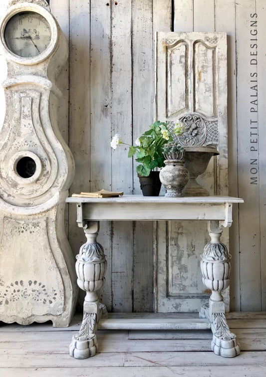 A hand-painted Versailles carved table with Annie Sloan Chalk Paint in a neutral color, featuring intricate leg designs, placed against a rustic wooden backdrop with decorative items on top.